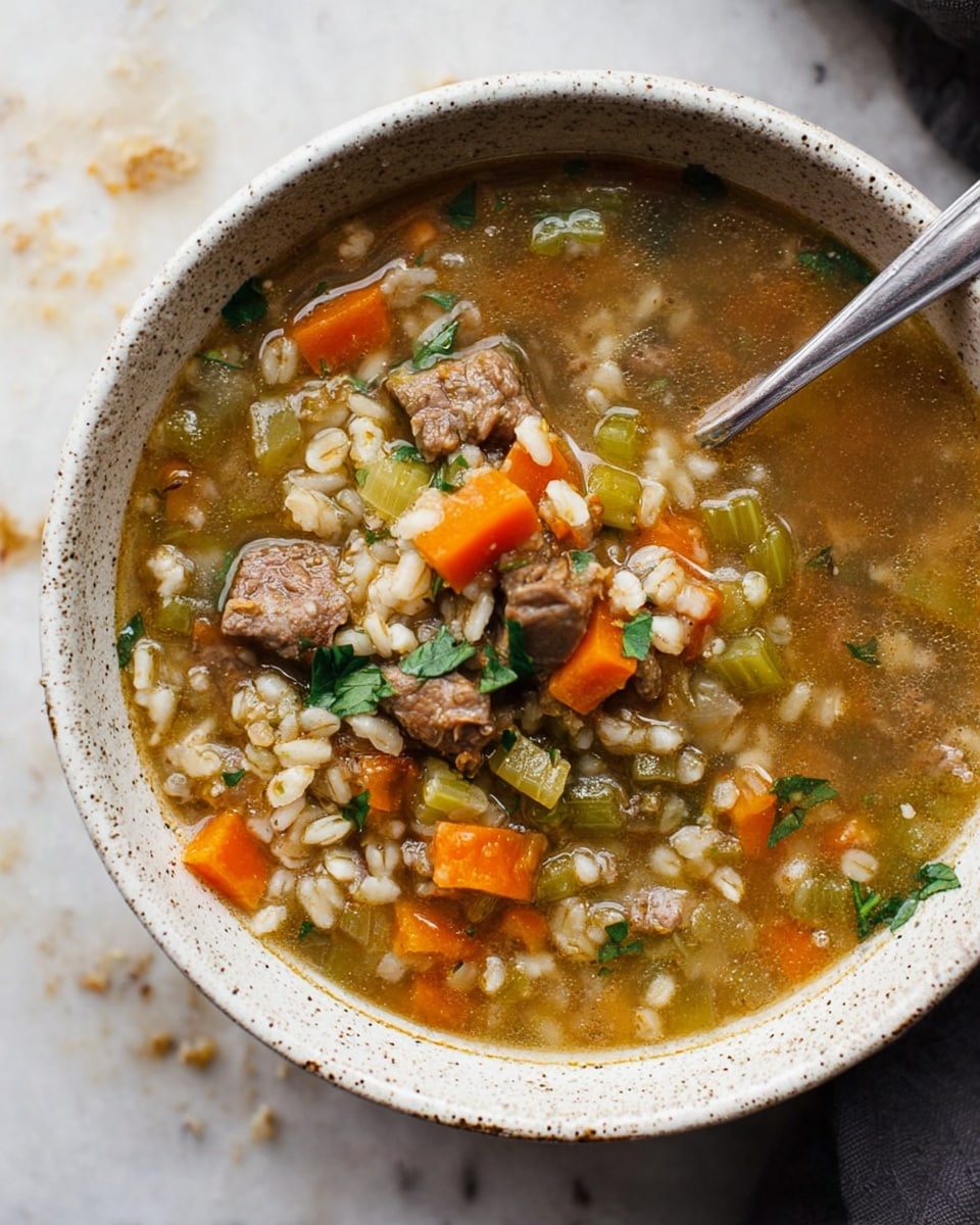 A white speckled bowl filled with a soup containing three main layers: a light brown broth base with visible grains of barley, bright orange diced carrots, soft green celery pieces, and medium-sized chunks of brown cooked meat scattered evenly throughout. Fresh green herb bits are sprinkled on top, adding a touch of freshness. A silver spoon rests inside the bowl to the right, partially submerged in the soup. The bowl sits on a white marbled surface with some crumbs around it. The overall look is warm and hearty, showing a well-mixed and colorful texture. Photo taken with an iphone --ar 4:5 --v 7