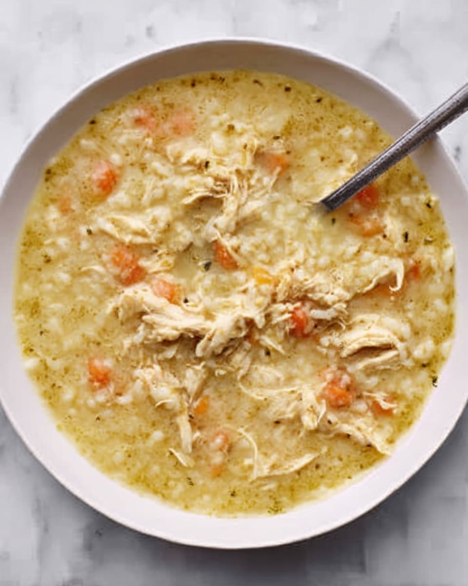 A white bowl filled with creamy chicken soup, showing shredded chicken pieces spread evenly throughout. The soup has a light yellowish color with small bits of carrot and rice visible inside. A spoon is partially dipped into the soup inside the bowl. The whole scene is set on a white marbled surface. Photo taken with an iphone --ar 4:5 --v 7