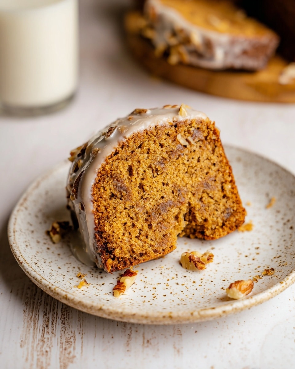 A single slice of moist, golden-brown spiced cake sits on a white speckled plate, showing a crumbly texture with small chunks of nuts embedded throughout. The cake has a thin layer of light, slightly shiny white glaze around its sides, with whole nut pieces peeking through the glaze unevenly. A small piece is taken out from one edge, revealing its soft interior. The plate rests on a surface with a white marbled texture, and blurred pieces of the whole cake and a glass of milk are faintly visible in the background. Photo taken with an iphone --ar 4:5 --v 7