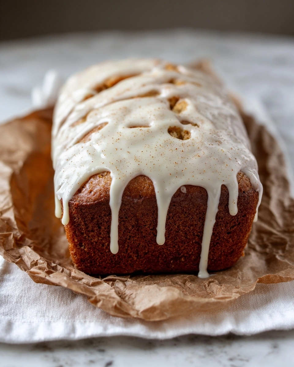 A loaf cake with a rich golden brown color sits on a piece of crumpled brown parchment paper, resting on a white cloth on a white marbled surface. The cake is topped with soft white icing that drips down the sides in thick, uneven streams, with the icing surface showing a few small holes and a sprinkle of light brown spice powder. The loaf has a soft and slightly cracked texture under the icing. Photo taken with an iphone --ar 4:5 --v 7