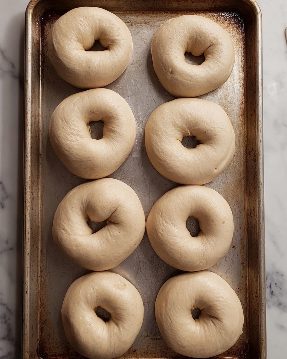 The image shows a baking tray filled with twelve uncooked bagels arranged in four rows of three. Each bagel is thick, pale beige in color, smooth in texture with some slight creases, and shaped into a rounded ring with a hole in the center. The bagels rest directly on a metal tray that has some scratches and slight discoloration from use. The background is a white marbled texture. photo taken with an iphone --ar 4:5 --v 7