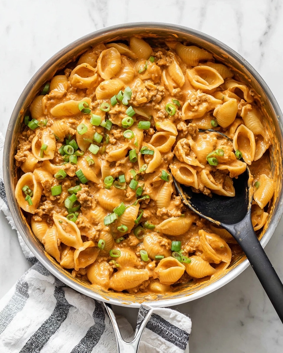 A silver pan filled with one layer of shell pasta coated in a thick creamy orange sauce, mixed with small bits of brown ground meat, topped with chopped bright green onions scattered evenly across the surface. A black spoon is scooping some pasta from the pan. The setting is on a white marbled texture with a white and black striped cloth partly visible under the pan. Photo taken with an iphone --ar 4:5 --v 7