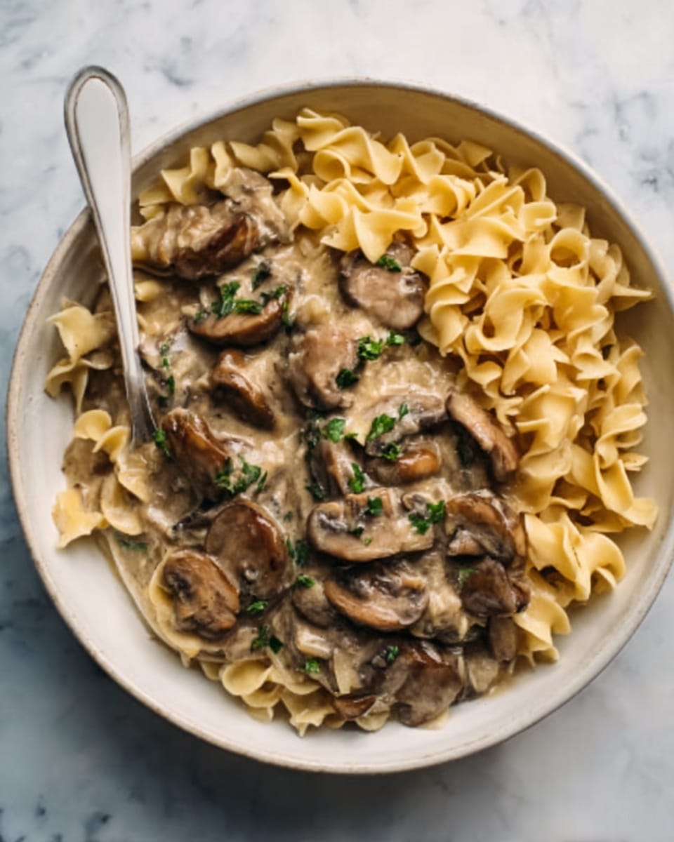 The image shows a white bowl filled with twisted egg noodles on one side and creamy mushroom sauce on the other. The creamy sauce has many light brown sliced mushrooms mixed in, with small green herb pieces sprinkled on top. A white spoon is placed inside the bowl, resting on the mushrooms. The bowl sits on a white marbled surface. Photo taken with an iphone --ar 4:5 --v 7
