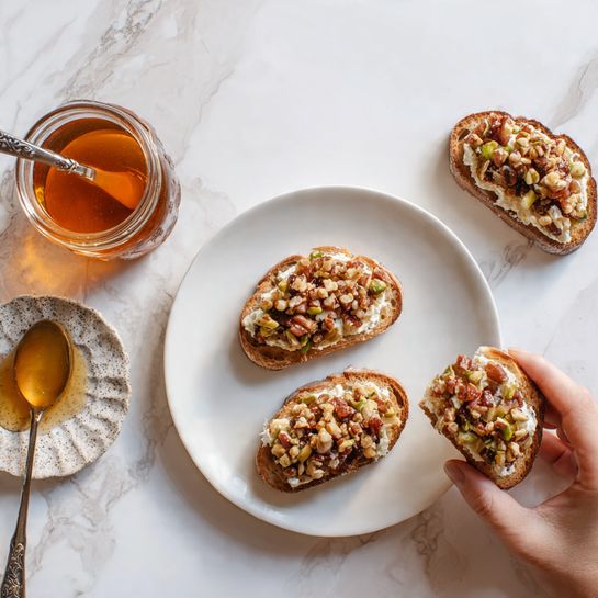 The image shows three toasted pieces of bread on a white marbled surface, each topped with a layer of creamy white spread and then a thick layer of finely chopped reddish-brown mixture. To the top left, there is a small glass jar filled with amber-colored honey, and beside it a white plate holding a silver spoon with honey dripping off it. A woman's hand is holding one of the toasted breads on the right side. The overall colors are warm with a mix of white, brown, and honey gold tones. Photo taken with an iphone --ar 4:5 --v 7