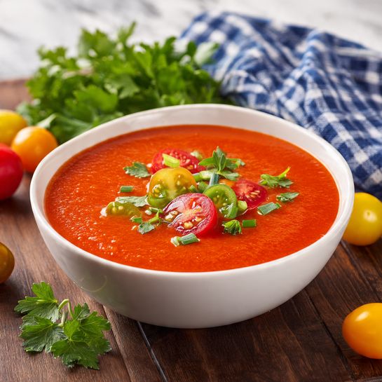 A white bowl filled with smooth, bright red tomato soup sits center on a dark wooden surface. The soup is topped with small slices of red and green cherry tomatoes and tiny pieces of green herbs. Around the bowl, there are whole and sliced red, yellow, and green cherry tomatoes scattered along with fresh green parsley leaves. A blue and white checkered cloth and more parsley leaves are placed behind the bowl. The background is a white marbled texture. Photo taken with an iphone --ar 4:5 --v 7