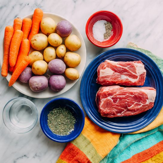 The image shows two white plates on a white marbled surface. The plate on the left holds baby carrots that are bright orange and round small potatoes that are light purple, filling the plate evenly. The plate on the right shows a thick raw piece of red meat with white marbling, lying flat. In front of the plates, there are three small round bowls: one blue bowl with clear water, one red bowl containing salt and pepper, and one green bowl filled with mixed green herbs or spices. A colorful cloth with yellow, orange, and green stripes is placed partially under the right plate. Photo taken with an iphone --ar 4:5 --v 7