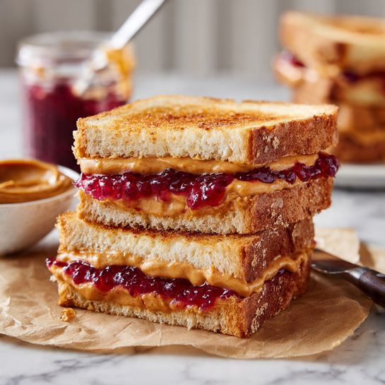 The image shows two toasted sandwich halves stacked on each other on a piece of brown paper. The sandwich has three slices of golden-brown toasted bread with a crispy texture. Between the layers, there is a spread of soft light orange peanut butter and bright red jelly with a glossy, smooth look. The sandwich is on a white marbled surface with a blurred white bowl of jelly and an open jar of peanut butter in the background. A woman's hand holds a knife inside the peanut butter jar. The photo taken with an iphone --ar 4:5 --v 7