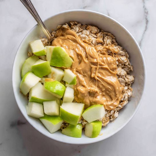 A white bowl sits on a white marbled surface, filled with a base layer of light brown oats with a rough texture. On top, there is a creamy, smooth layer of light brown peanut butter sauce spread unevenly, covering most of the oats. Scattered on the peanut butter are several chunks of bright green apple with white flesh, some coated in the peanut butter sauce. A silver spoon is partially visible, resting inside the bowl on the left side. Photo taken with an iphone --ar 4:5 --v 7