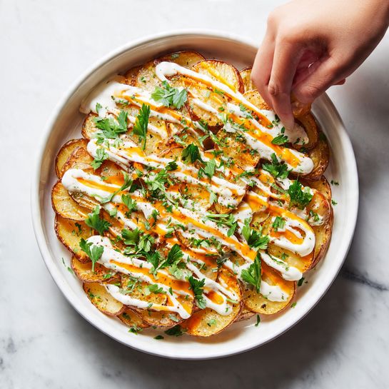 A white bowl filled with roasted golden-brown potato pieces arranged in layers, topped with drizzles of creamy white sauce and bright orange sauce that adds a smooth texture. Fresh green parsley leaves are scattered on top, adding a touch of color. A woman's hand is seen reaching in to drizzle or sprinkle over the dish. The bowl sits on a white marbled surface. Photo taken with an iphone --ar 4:5 --v 7