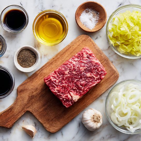 A wooden board holds a fresh raw marbled beef chunk with red and white textures on the upper right. Surrounding it are small clear round bowls: one with light golden oil, one with sliced pale yellow-green peppers, and one with finely sliced white onions in the lower right. To the left are small glass dishes with black pepper, salt, dark soy sauce, and another dark brown liquid. A small bulb of garlic is near the lower edge of the board. All items are placed on a white marbled surface. photo taken with an iphone --ar 4:5 --v 7