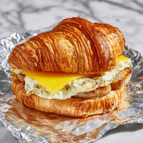 The image shows a croissant sandwich placed on shiny foil with a white marbled texture underneath. The croissant is golden brown and flaky, topped with a layer of melted yellow cheese visible through an opening. Below the cheese, there is a piece of cooked sausage with a browned, slightly shiny surface. Bitter-sweet and slightly crispy fried egg is layered above the sausage, with uneven white edges. The sandwich looks warm and freshly made. Photo taken with an iphone --ar 4:5 --v 7