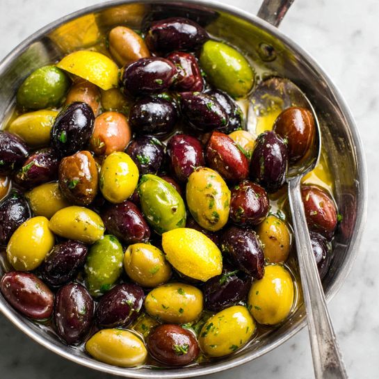 This image shows a close-up of a pile of green olives placed in the middle of a white plate. The olives are shiny and covered with small pieces of herbs and spices in black, red, and green colors. They sit in a thin layer of golden olive oil that pools around the base of the olives. The olives vary slightly in size, and their smooth, round shapes fill the plate. The plate rests on a white marbled surface, visible faintly at the edges. photo taken with an iphone --ar 4:5 --v 7