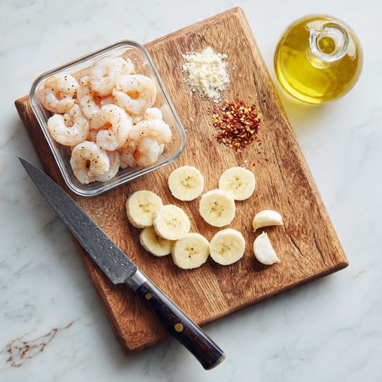 The image shows a wooden cutting board placed on a white marbled surface. On the cutting board, there are thin banana slices arranged in a small pile at the center-right side, a few small garlic cloves lined up near the top-right corner, and some salt and crushed red pepper spice in small piles near the top center. To the left of the cutting board, there is a clear square glass container filled with peeled, raw shrimp. A large knife with a black handle rests diagonally on the bottom left corner of the cutting board. In the top right corner of the image, there is a glass bottle of olive oil. The overall scene looks like a preparation area for cooking. Photo taken with an iphone --ar 4:5 --v 7