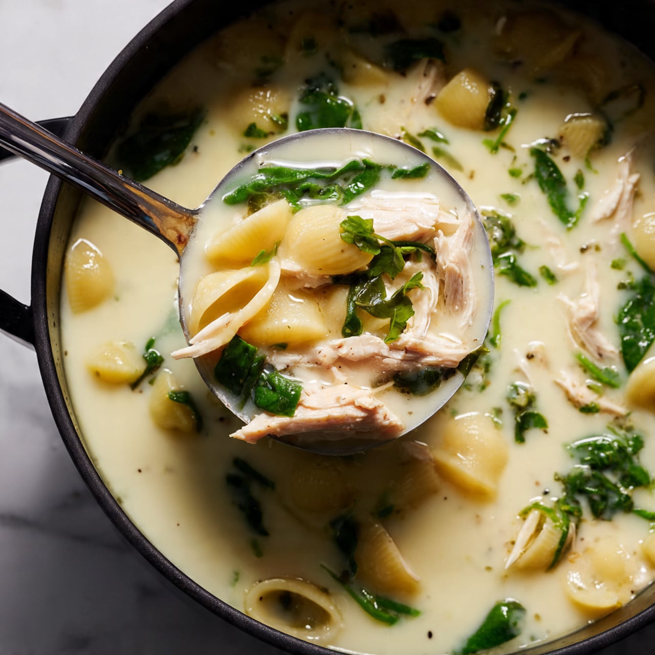 A close-up of a ladle holding creamy white soup with visible layers: shredded light beige chicken pieces, soft pale yellow pasta shells, and bright green spinach leaves, all immersed in a smooth, milky broth. The ladle is silver and sits inside a black pot filled with the same soup, placed on a white marbled surface. The image captures the texture of the veggies and chicken floating in the liquid, highlighting the mix of creamy and fresh elements. photo taken with an iphone --ar 4:5 --v 7