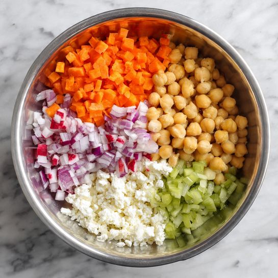 A silver bowl with six different ingredients placed separately inside: bright orange diced carrots at the top right, pale yellow chickpeas to the right below the carrots, light green chopped celery at the bottom, white crumbled cheese in the middle, small red and white diced radishes on the left below the chopped red onions, which are at the upper left. The bowl rests on a white marbled surface. photo taken with an iphone --ar 4:5 --v 7