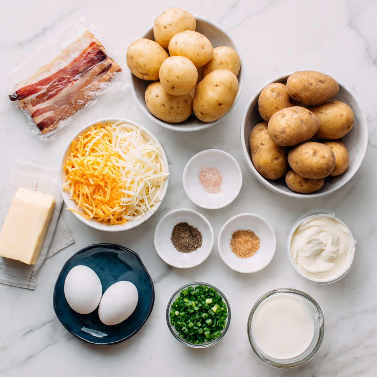 The image shows ingredients laid out neatly on a white marbled surface. At the top center, there is a white bowl filled with six smooth light brown potatoes. To the right, another white bowl contains five darker brown potatoes. On the left side, there is a package of uncured beef bacon partly visible. Below it, a large white bowl holds three layers of shredded cheese in colors of orange, white, and pale yellow, stacked from left to right. Near the center, there are three small white plates; one holds black and light brown powders, the second has a pinkish salt, and the third is filled with chopped green chives. A small glass cup next to them contains white cream. Below this setup, a dark blue small plate carries two white eggs. To the right, there is a clear measuring cup with a white liquid inside, and above it, a stick of butter wrapped in paper rests horizontally. Photo taken with an iphone --ar 4:5 --v 7
