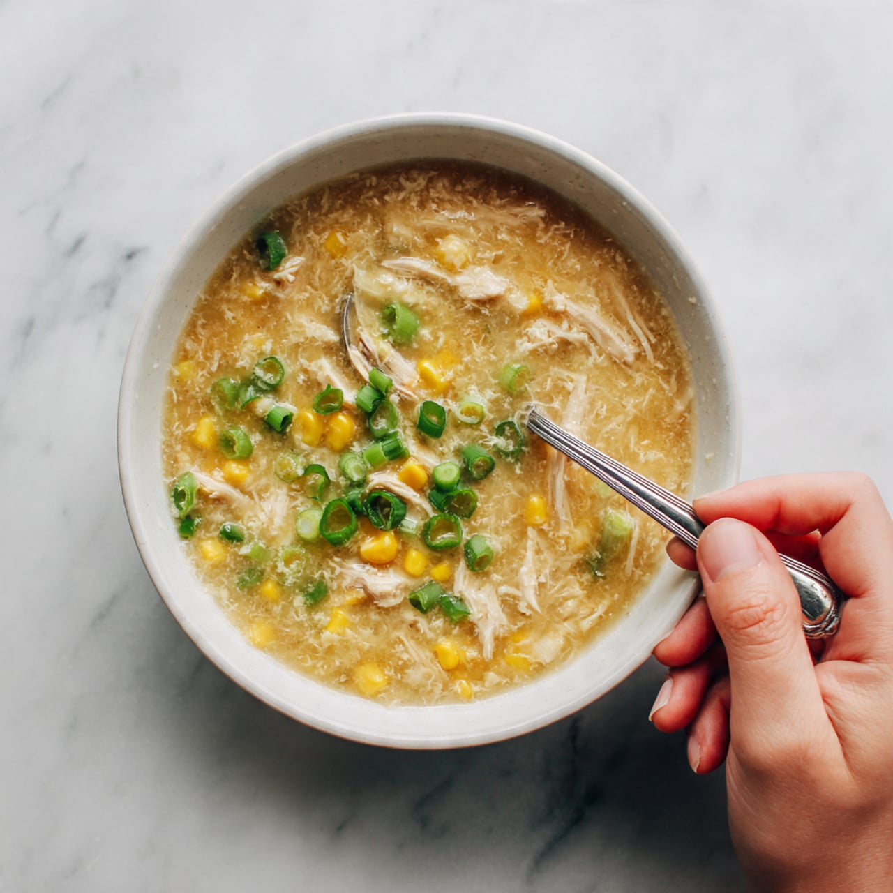 A white bowl filled with thick soup sits on a white marbled surface, the soup showing a pale golden color with visible pieces of shredded chicken, bright yellow corn kernels, and finely chopped green onions scattered on top as garnish; a silver spoon rests inside the bowl with a woman's hand holding it gently from the right side. Photo taken with an iphone --ar 4:5 --v 7