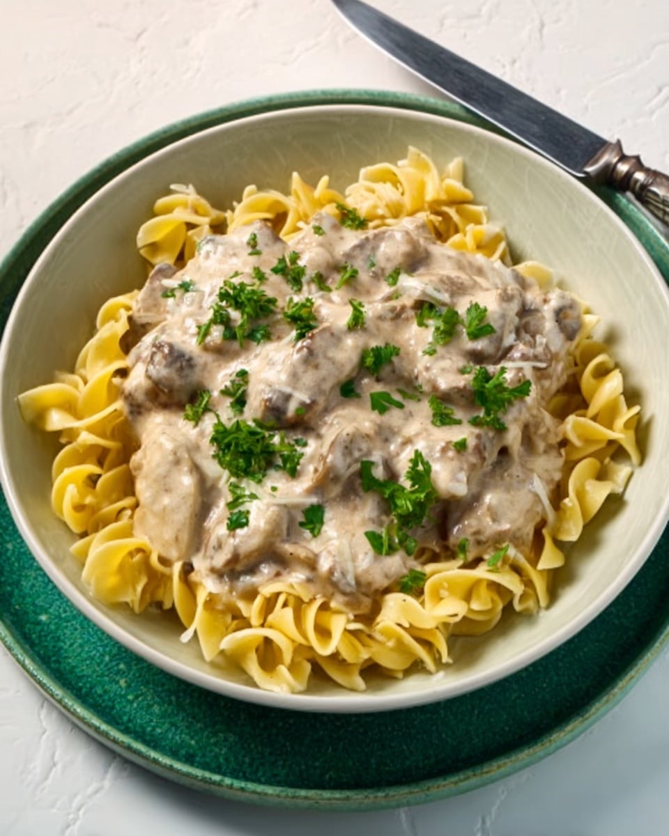 A white bowl filled with yellow curly egg noodles at the bottom, topped with a thick layer of creamy light beige sauce mixed with small pieces of tender meat. Fresh green parsley leaves are sprinkled on top as garnish. The bowl sits on a green chalky plate, and a knife is placed next to the plate on a white marbled surface. Photo taken with an iphone --ar 4:5 --v 7