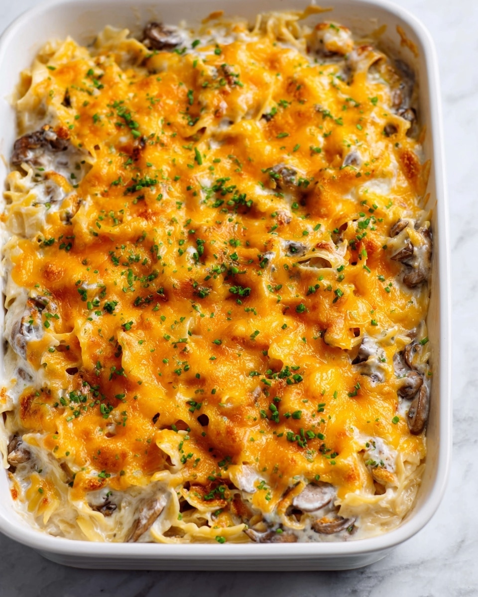 A white rectangular baking dish filled with layers of cooked pasta mixed with a creamy white sauce and small pieces of mushrooms throughout. The top layer is covered with melted bright orange cheddar cheese that is slightly bubbling and stretchy in texture, sprinkled with small green chopped herbs for color contrast. The dish looks warm and inviting, sitting on a white marbled surface. photo taken with an iphone --ar 4:5 --v 7
