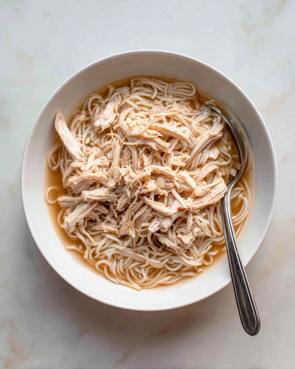 The image shows a white bowl filled with a dish that has shredded light brown chicken pieces mixed with thin white noodles. The dish is covered with a light brown sauce that makes the noodles and chicken look glossy and wet. There is a silver spoon placed inside the bowl on the right side, slightly buried in the food. The bowl sits on a white marbled surface, and the photo was taken with an iphone --ar 4:5 --v 7