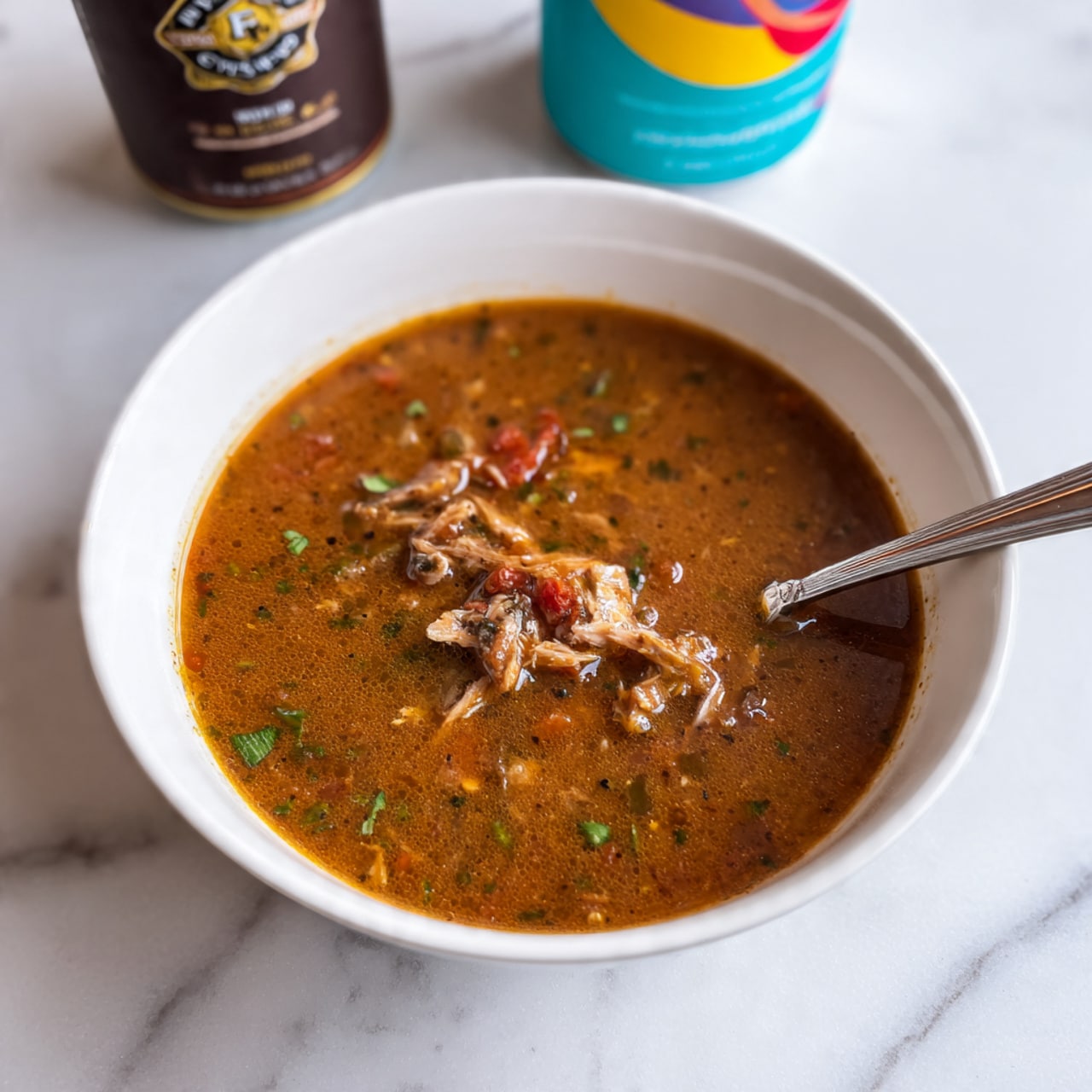 The image shows a white bowl filled with a brown soup that has chunks of meat and small pieces of green herbs floating in it. The soup looks thick and hearty, with a few pieces of shredded meat visible throughout. A silver spoon is inside the bowl on the right side, partially submerged in the soup. The bowl sits on a white marbled surface, and behind it are two cans: one dark brown with a label, and the other light blue and yellow with a colorful design. The scene is simple and focused on the bowl of soup. photo taken with an iphone --ar 4:5 --v 7