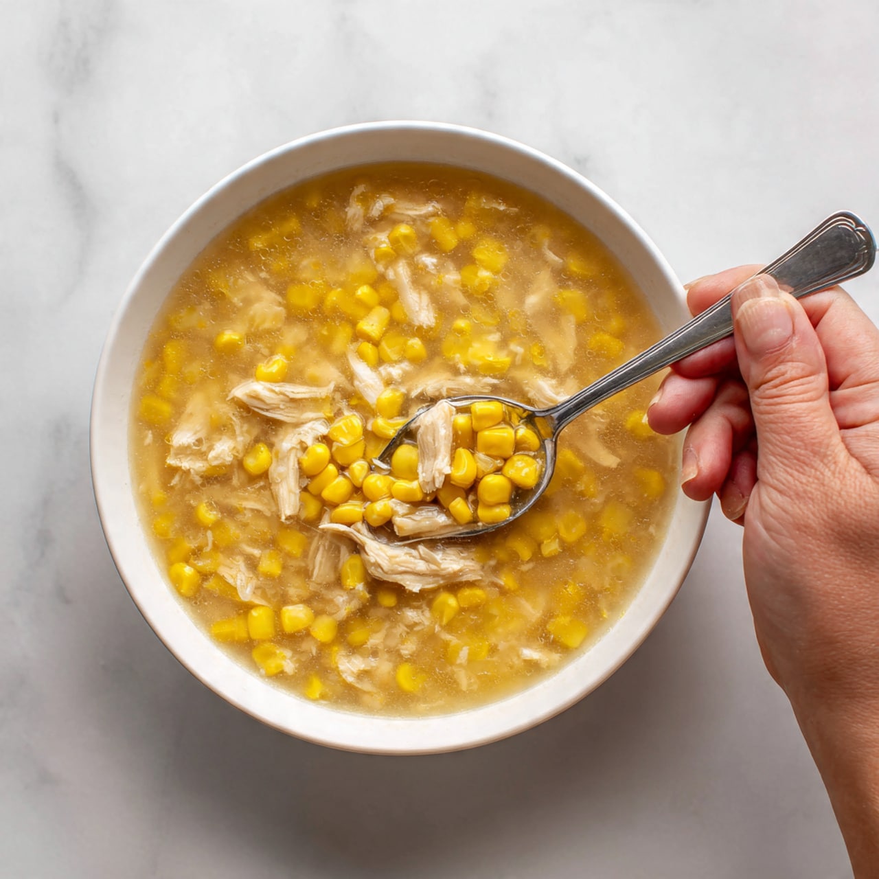The image shows a white bowl filled with a clear broth containing yellow corn kernels and pieces of light-colored chicken. The soup looks warm and hearty, with the corn and chicken evenly mixed in the broth. A silver spoon is placed inside the bowl, with a woman's hand holding the spoon from the top right corner of the image. The bowl sits on a white marbled surface, highlighting the light colors of the soup. Photo taken with an iphone --ar 4:5 --v 7