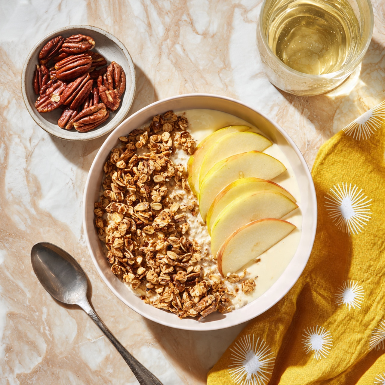 A white bowl filled with creamy oatmeal is topped with a layer of chunky, brown granola covering most of the surface. On one side, three thin slices of pale yellow apple with smooth edges are placed neatly in a fan shape on top of the granola. Next to the bowl is a small white bowl containing shiny, dark brown pecans. The setting includes a white marbled surface beneath the bowl and a yellow cloth with white sun patterns nearby. A silver spoon and a glass of light-colored liquid are also visible. A woman's hand slightly touches the yellow cloth near the bowl. Photo taken with an iphone --ar 4:5 --v 7