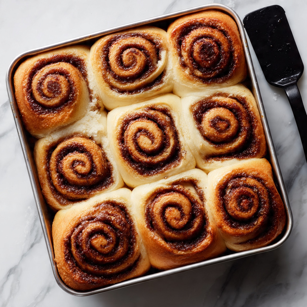A silver baking pan filled with nine cinnamon rolls arranged in three rows and three columns. Each cinnamon roll has golden-brown, slightly shiny tops with visible dark brown cinnamon swirls spiraled inside soft, fluffy layers. The rolls are tightly packed, with some edges touching, showing light, fluffy sides with a slight fluffiness texture. The pan is placed on a white marbled surface next to a black cooking spatula with a smooth, matte finish. The image is taken from above, showing the rolls close-up. photo taken with an iphone --ar 4:5 --v 7