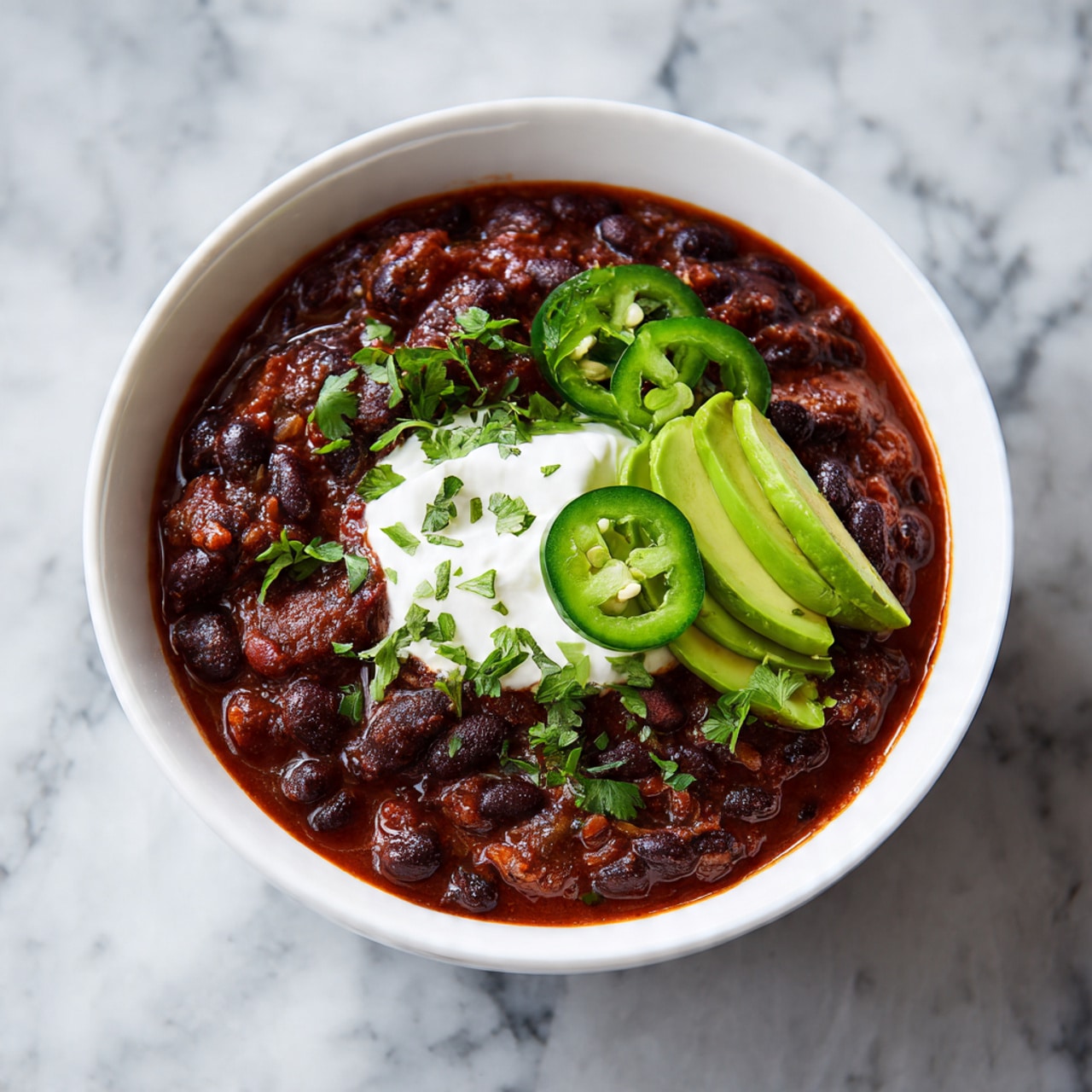 A white bowl filled with dark brown and black beans in a thick red sauce topped with a dollop of white sour cream in the center. On one side are three fresh green slices of avocado, and on top of the sour cream are two green round slices of jalapeño and small green herb leaves scattered on the surface. The bowl sits on a white marbled surface. photo taken with an iphone --ar 4:5 --v 7