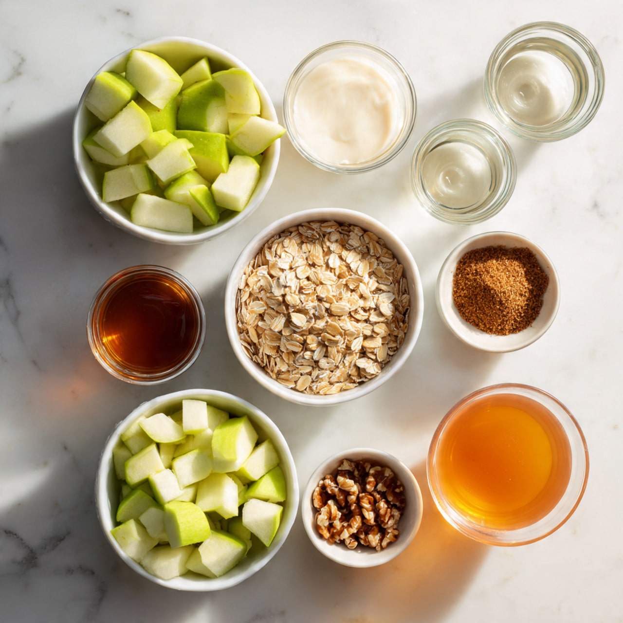 The image shows a white marbled surface with several small white bowls and glass containers arranged neatly. There are small white bowls filled with light green chopped apples, light brown rolled oats, and dark brown chopped nuts. A glass container holds a deep orange liquid, and another has a white creamy liquid. Other small white bowls contain vanilla extract and a brown powdery spice. Two clear small glasses are empty, placed side by side. The whole scene is bright and clean, organized in a simple layout. photo taken with an iphone --ar 4:5 --v 7