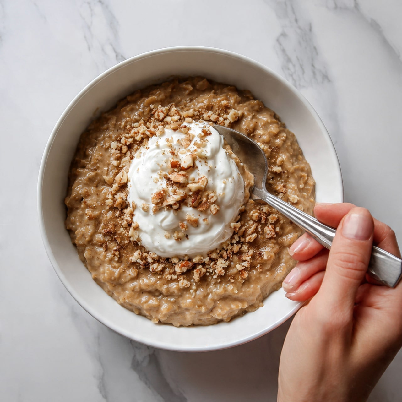 A white bowl filled with thick, brown oatmeal that looks creamy and warm. On top of the oatmeal, there is a sprinkle of light brown crushed nuts or crumbs covering most of the surface. In the center, there is a dollop of white whipped cream or yogurt with a soft, fluffy texture. A spoon is placed inside the bowl, with a woman's hand holding it from the side. The bowl is set on a white marbled surface. photo taken with an iphone --ar 4:5 --v 7