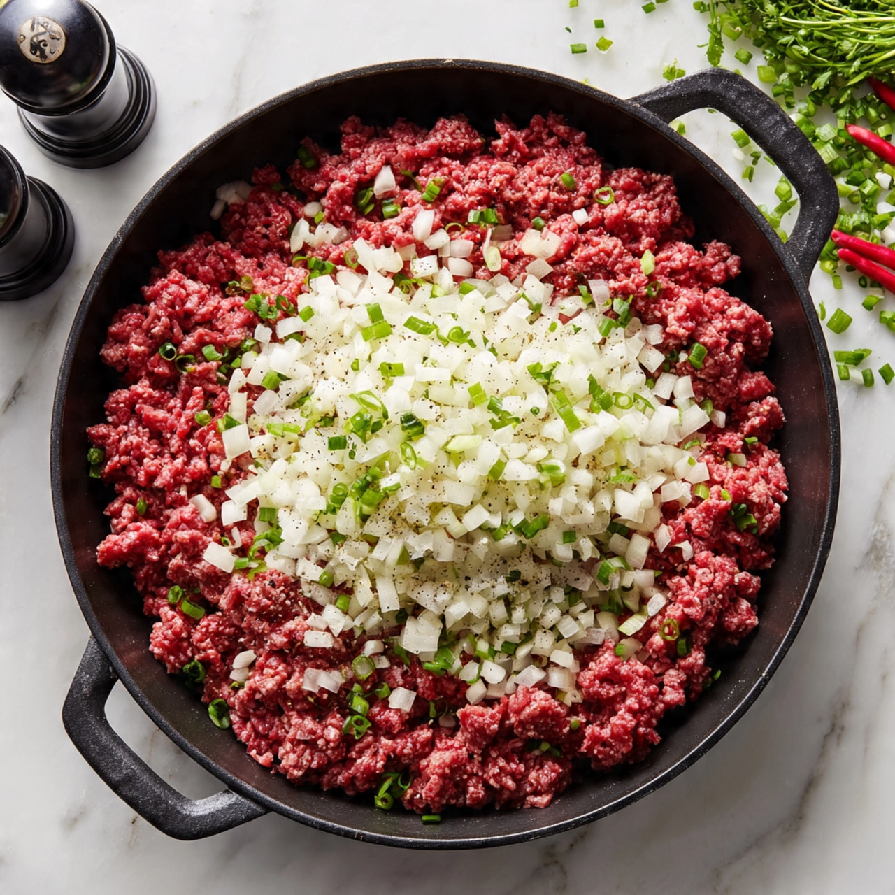 A black skillet filled with raw ground beef forming the base layer, topped with a layer of small chopped white onions scattered mostly in the center. The skillet handles are visible at the top and bottom edges. The background is a white marbled surface with two small pepper grinders and some green and red garnishes partially visible at the top right. Photo taken with an iphone --ar 4:5 --v 7