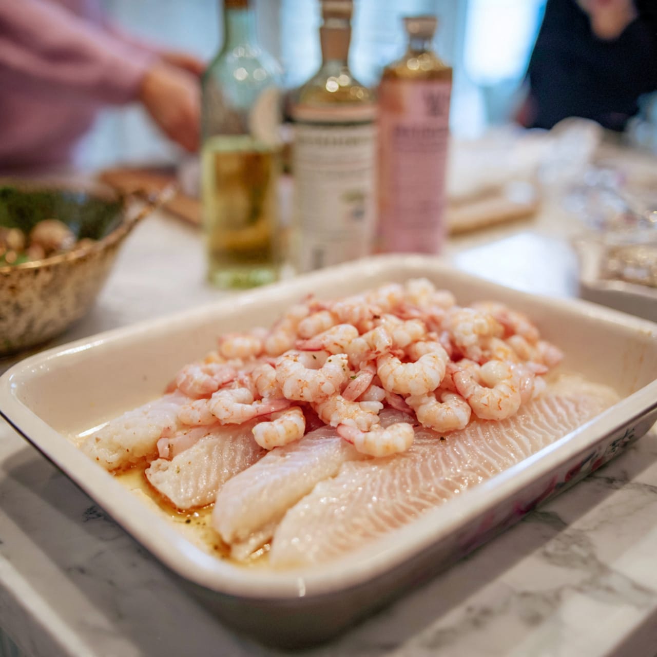 The image shows a white pan with two thick, pale fish fillets at the bottom, topped with a large pile of small, light pink raw shrimp scattered unevenly over the fish. The background includes two bottles that are partially out of focus on a white marbled surface. A woman's hand is not visible here. Photo taken with an iphone --ar 4:5 --v 7