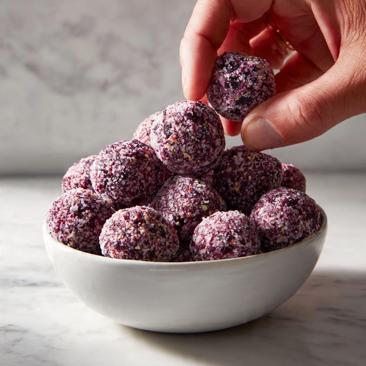 A close-up view of a large white bowl filled with many small round bite-sized balls coated in a thick, light purple layer with darker purple specks inside each ball. The balls have a rough and slightly powdery texture. A man's hand is reaching into the bowl, pinching one ball between the thumb and forefinger. The bowl sits on a white marbled surface with subtle patterns. The image is well-lit with natural light highlighting the texture and colors of the balls. photo taken with an iphone --ar 4:5 --v 7