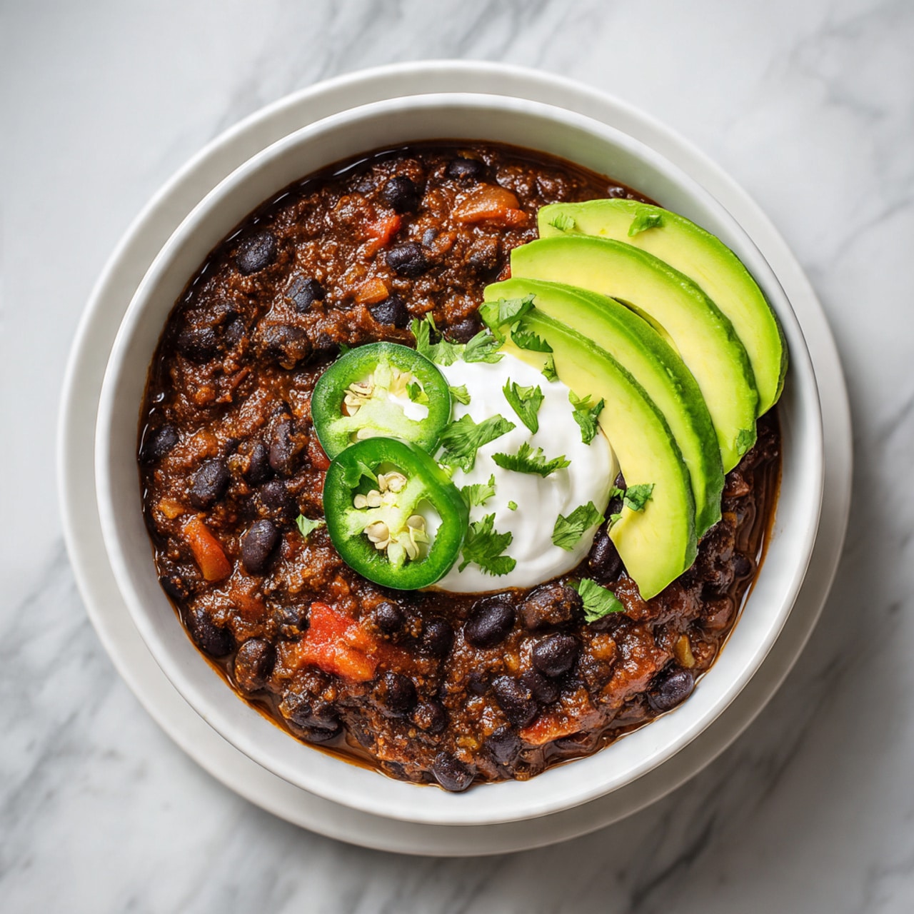 The image shows a bowl filled with black bean stew, with a dark brown, thick and chunky texture made up of black beans and bits of tomato. On top, there is a layer of white sour cream placed near the center, with two green jalapeño slices resting on it. Next to the sour cream, there are several slices of bright green avocado arranged neatly. The bowl itself is white with a simple design, placed on a white marbled surface. Photo taken with an iphone --ar 4:5 --v 7