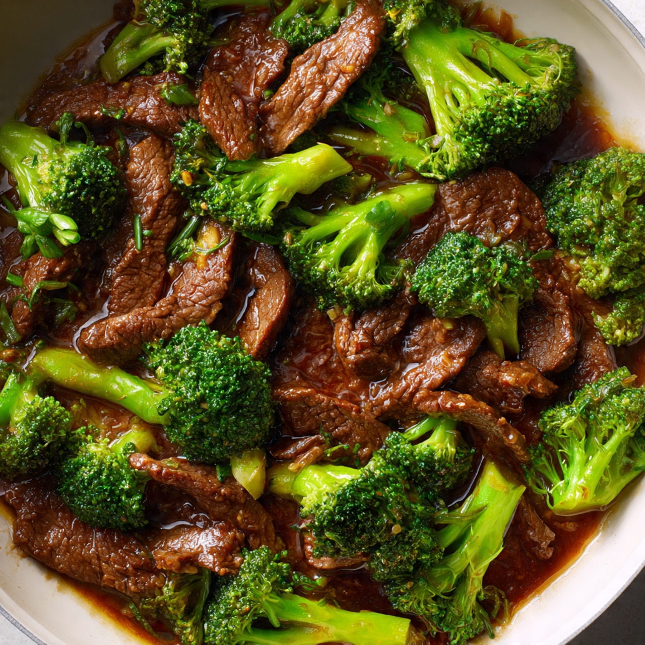 The dish shows cooked sliced beef strips that are dark brown and tender, mixed with bright green broccoli florets. The broccoli is fresh and slightly steamed, scattered evenly among the beef. The beef pieces have a shiny texture from a rich, thick brown sauce that coats both the meat and broccoli, pooling slightly at the bottom of the white pan. The image gives a close-up look inside the pan, showing a mix of colors from dark brown sauce, green broccoli, and browned beef all combined together. Photo taken with an iphone --ar 4:5 --v 7