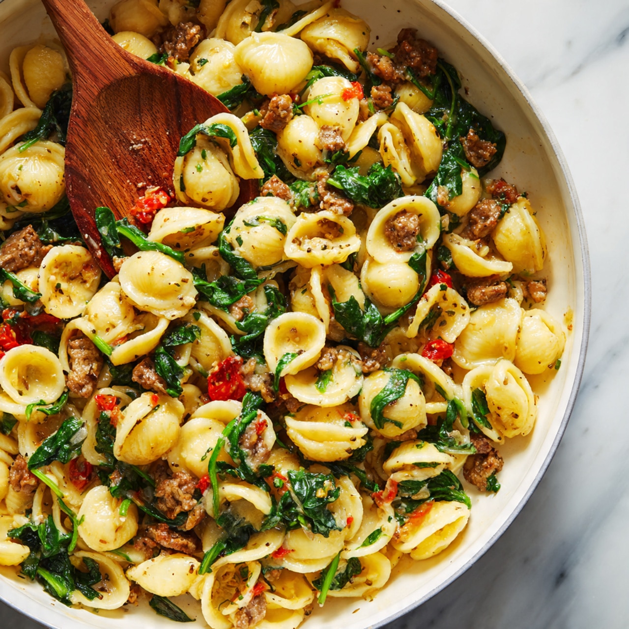 A close-up view of a white pan filled with cooked orecchiette pasta mixed with small pieces of browned meat, wilted green leafy vegetables, and small red tomato pieces. A wooden spoon is partially buried in the pasta on the left side, lifting some of the mixture. The pasta pieces are light yellow with a slight shine, and the meat is a golden brown with some crispy edges. The green leaves add a fresh contrast, and the small red bits provide a pop of color throughout. The background is a white marbled surface. photo taken with an iphone --ar 4:5 --v 7