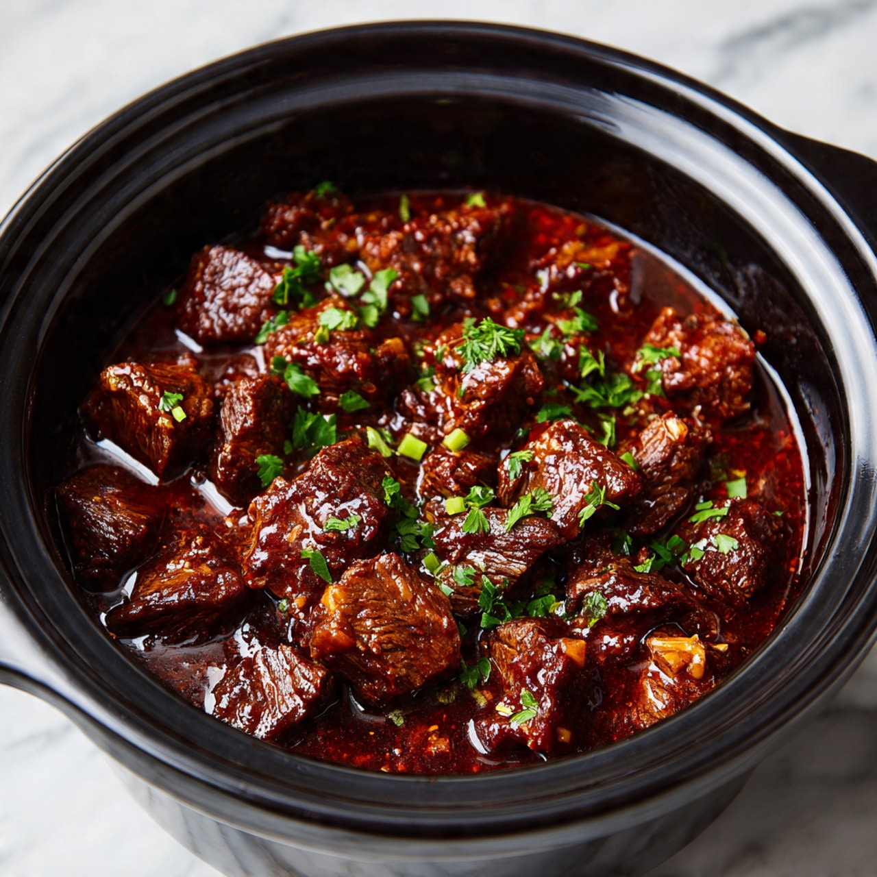 A close-up view of a black slow cooker filled with about three layers of dark brown beef chunks cooked in a thick, glossy reddish-brown sauce, topped with small pieces of chopped green herbs scattered evenly over the meat. The beef pieces vary slightly in size, with a tender, well-cooked texture and some visible strands of meat fibers. The sauce has a rich shine that coats the beef, pooling slightly at the bottom, and the inside of the cooker has a matte black finish. The photo is set against a white marbled surface. Photo taken with an iphone --ar 4:5 --v 7