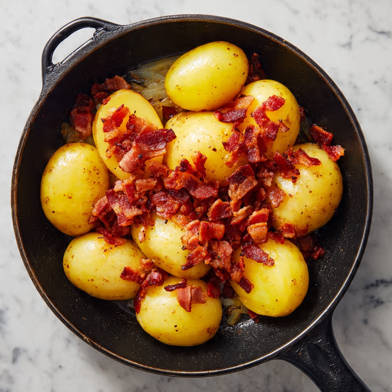 A black cast iron pan on a gas stove holds a dish with two main layers: the bottom layer is small cooked bacon pieces with a crispy texture and reddish-brown color, spread unevenly in the pan, and the top layer consists of several large yellow potato pieces, some whole and some halved, with smooth skin and a shiny finish. The colors contrast well with the dark pan and the white marbled surface around the stove. photo taken with an iphone --ar 4:5 --v 7