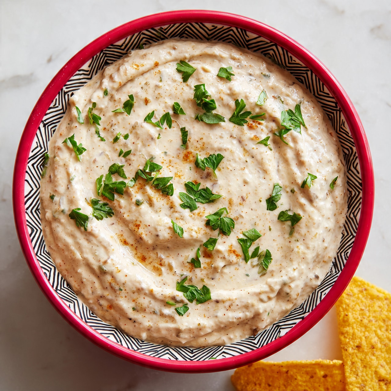 The image shows a white bowl filled with a creamy beige dip that has visible small chunks and a smooth but slightly lumpy texture. The dip is garnished with small green parsley leaves scattered on top, adding a fresh touch of color. The bowl has a bright red rim with a bold black and white geometric pattern, contrasting with the pale color of the dip. The bowl rests on a white marbled surface, and in the bottom right corner, there is a piece of yellow tortilla chip visible. photo taken with an iphone --ar 4:5 --v 7
