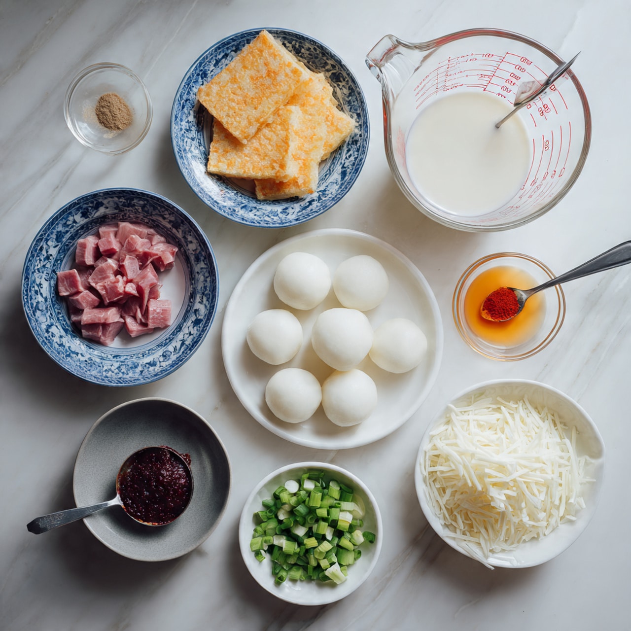 The image shows a variety of cooking ingredients arranged neatly on a white marbled surface. In the center, there is a white bowl filled with smooth, white rice cakes. Above it, a clear glass measuring cup filled with a milky white liquid sits with red measurement markings. To the top left, a white bowl with a blue pattern holds golden-brown crispy squares. To the bottom left, another white bowl with a blue pattern contains raw, pink meat pieces with a spoon resting inside. Below these bowls, a small gray bowl holds a dark red paste with a spoon. On the right side, a small white square bowl contains bright green chopped spring onions. Next to it, a tiny clear measuring cup has a small amount of orange liquid. Near the center bottom, a small white round bowl contains red powder, and to its right, a white square bowl is filled with shredded white cheese. All items are placed with some spacing, showing their textures and colors clearly, making a neat and organized cooking setup. Photo taken with an iphone --ar 4:5 --v 7