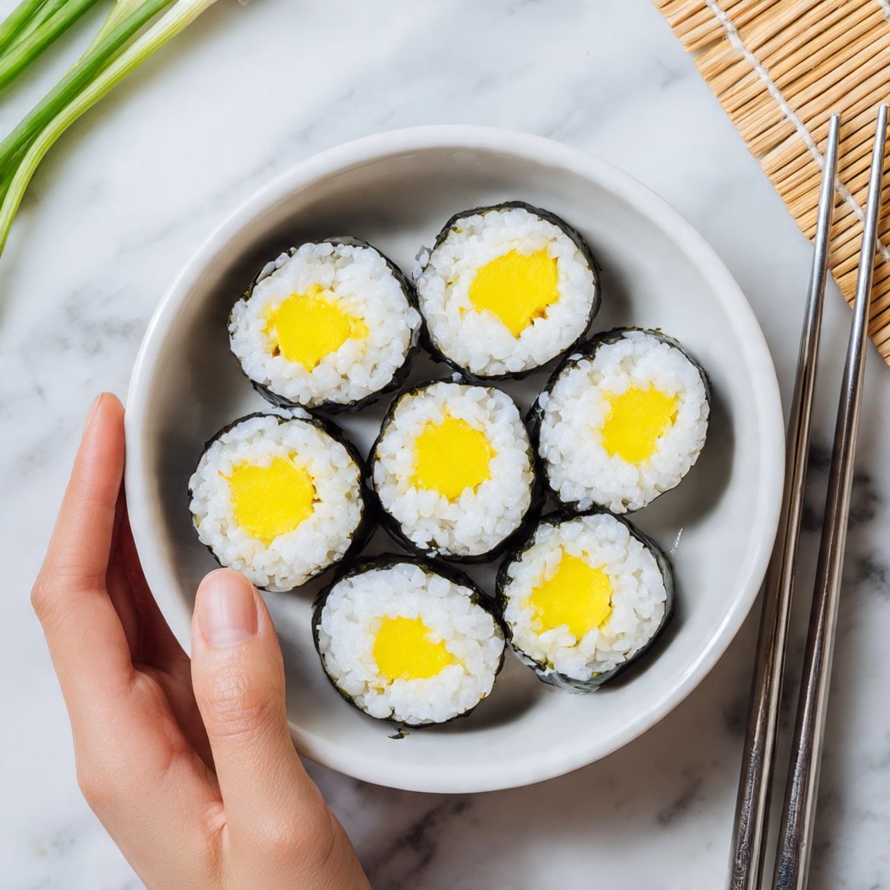 A white bowl filled with several pieces of sushi rolls is shown from above, placed on a white marbled surface with a beige bamboo mat underneath. Each sushi roll has three layers: a dark seaweed outer layer, a middle layer of white rice, and a bright yellow filling in the center that looks smooth and creamy. The pieces are arranged neatly in the bowl, showing round shapes and clean edges. A pair of silver chopsticks rests next to the bowl, with a woman's hand holding one piece of sushi at the top left. Green sliced onions are slightly visible at the top left of the image. Photo taken with an iphone --ar 4:5 --v 7