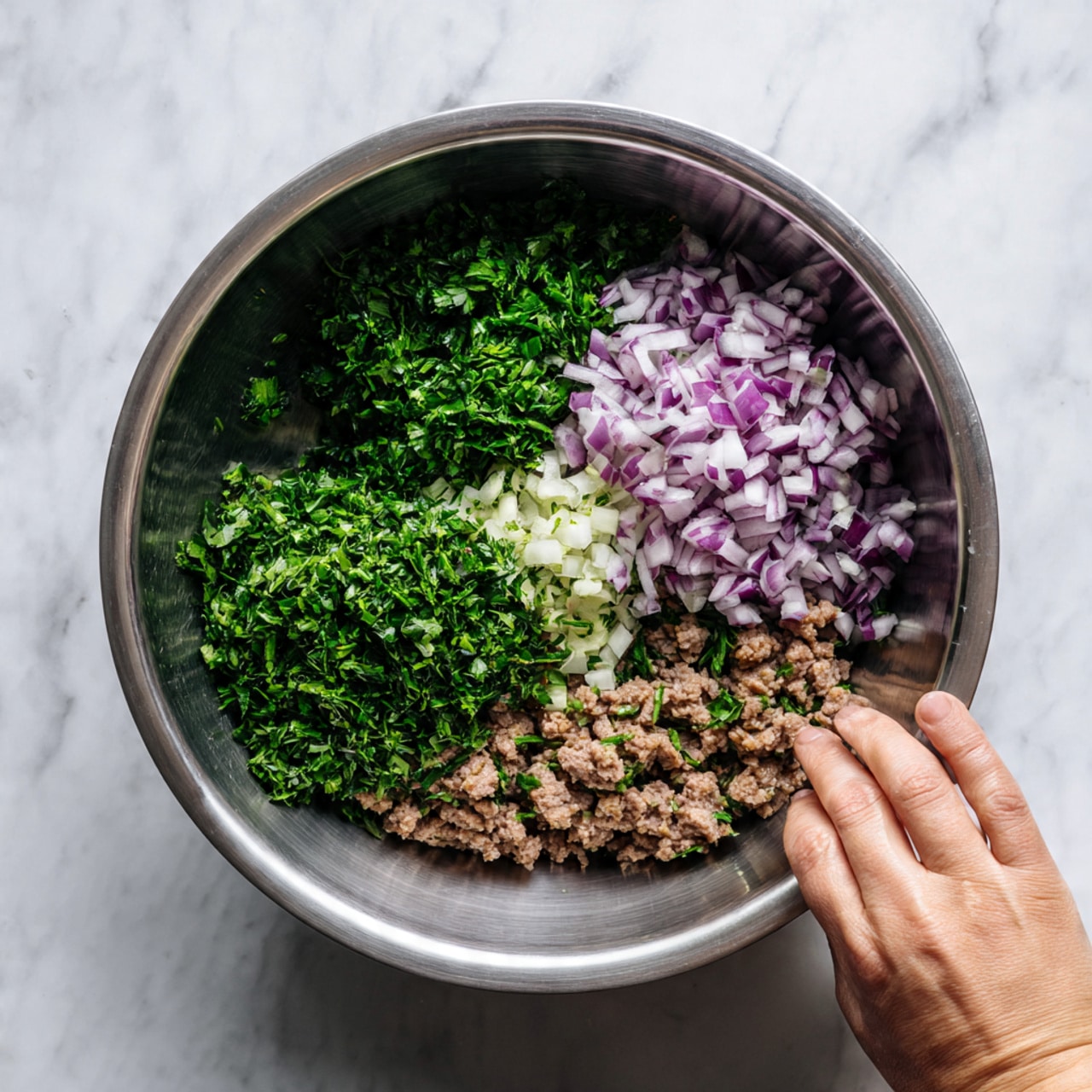 Inside a silver metal bowl, three separate layers sit side by side: finely chopped green herbs on the left with a rough texture, finely diced light purple onions on the right, and crumbled brown meat pieces at the bottom. The bowl shows a shiny reflection, and a woman's hand is touching the edge. The background is a white marbled texture. photo taken with an iphone --ar 4:5 --v 7