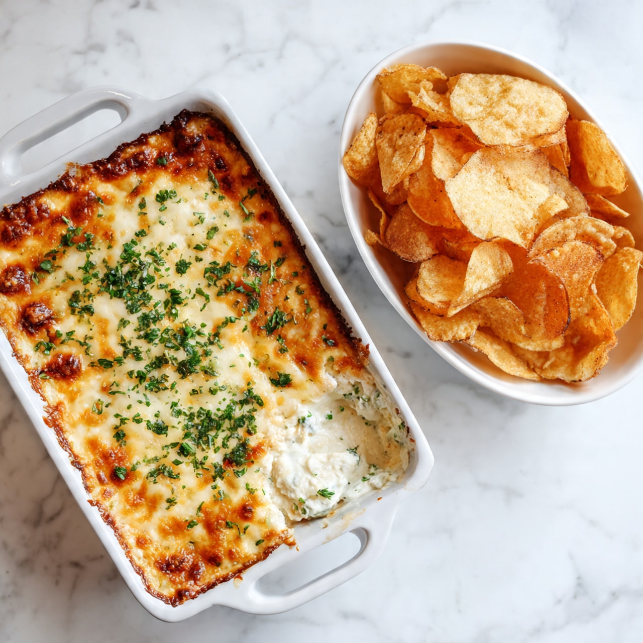 The image shows a white rectangular dish filled with a cheesy baked dip. The top layer is golden brown with melted cheese, sprinkled with chopped green herbs. The dip underneath is creamy and white, visible where a portion has been scooped out. Next to the dish, on a white marbled surface, is a white bowl filled with golden brown, crispy chips. The setting is bright and clean. Photo taken with an iphone --ar 4:5 --v 7