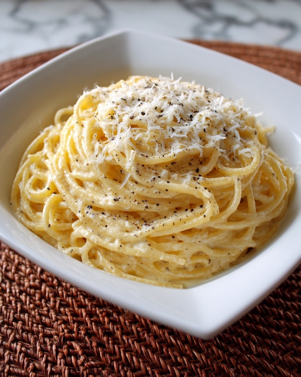 A round, thick pile of creamy spaghetti pasta sits in the center of a white square bowl. The pasta is coated in a smooth, light yellow sauce and topped with a layer of finely grated pale cheese. Small specks of black pepper sprinkle over the cheese, adding contrast. The bowl is placed on a brown woven mat, and the background is a white marbled surface. photo taken with an iphone --ar 4:5 --v 7
