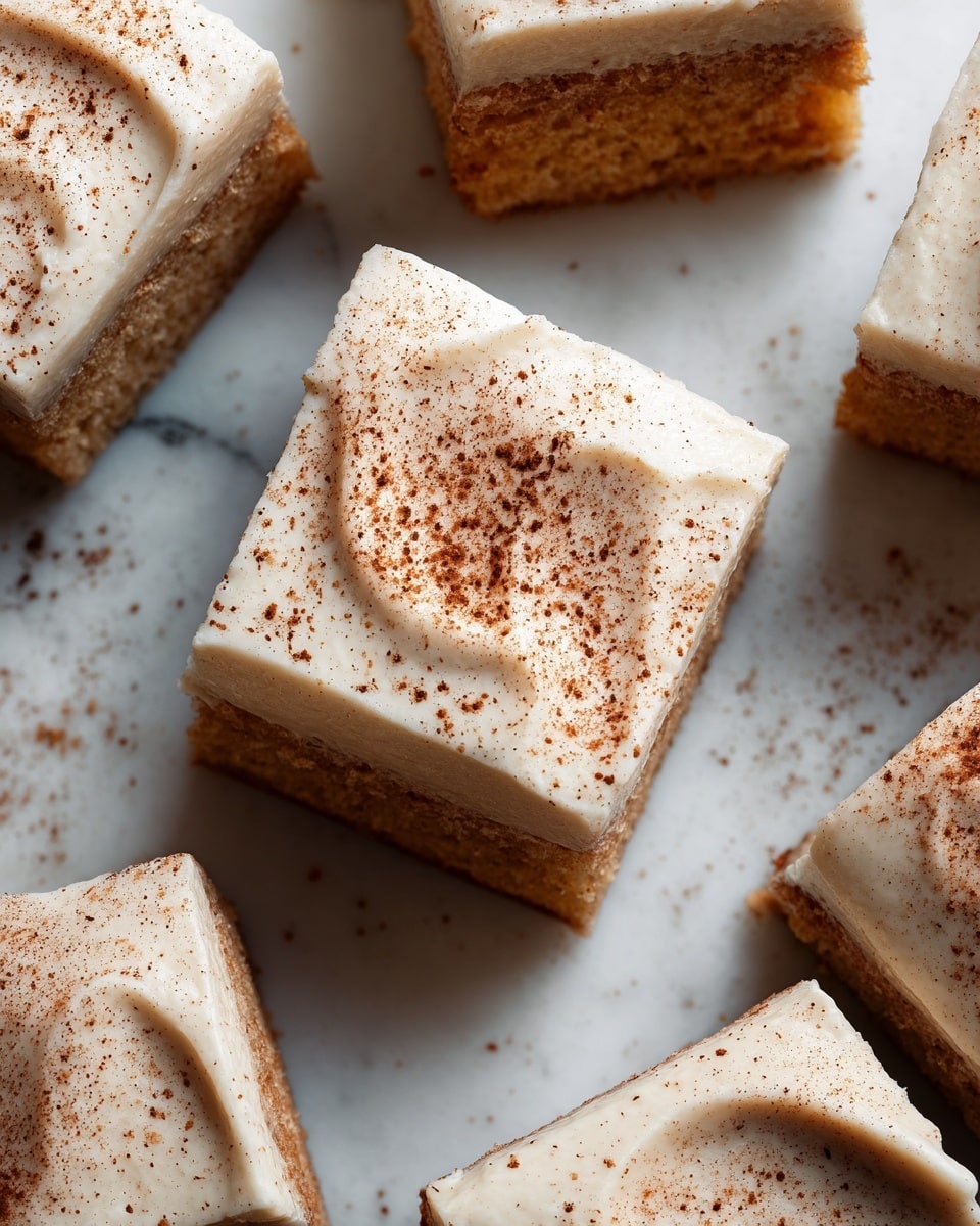 The image shows a close-up of several square pieces of moist cake with light beige frosting on top, each piece about one layer thick. The cakes have a soft, orange-brown color with a smooth creamy layer evenly spread above them. A light dusting of brown spice powder is sprinkled gently on the frosting surface. The texture of the cake looks soft and slightly crumbly, while the frosting is smooth and thick. The background is a white marbled texture. Photo taken with an iphone --ar 4:5 --v 7