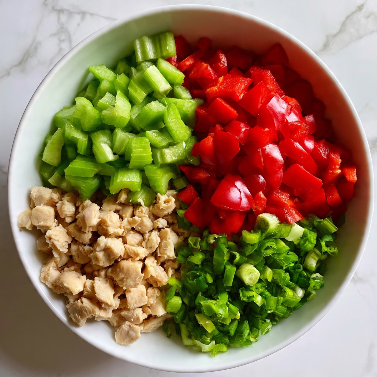 square image A white bowl is placed on a white marbled surface, filled with four different chopped ingredients neatly separated in sections. One section has small pieces of cooked chicken, slightly beige with a soft texture. Next to it, bright green celery chunks with a crisp, fresh look. Another section shows vibrant red bell pepper pieces, smooth and shiny. The last section contains chopped green onions, finely cut with a slightly curved shape. photo taken with an iphone --ar 4:5 --v 7