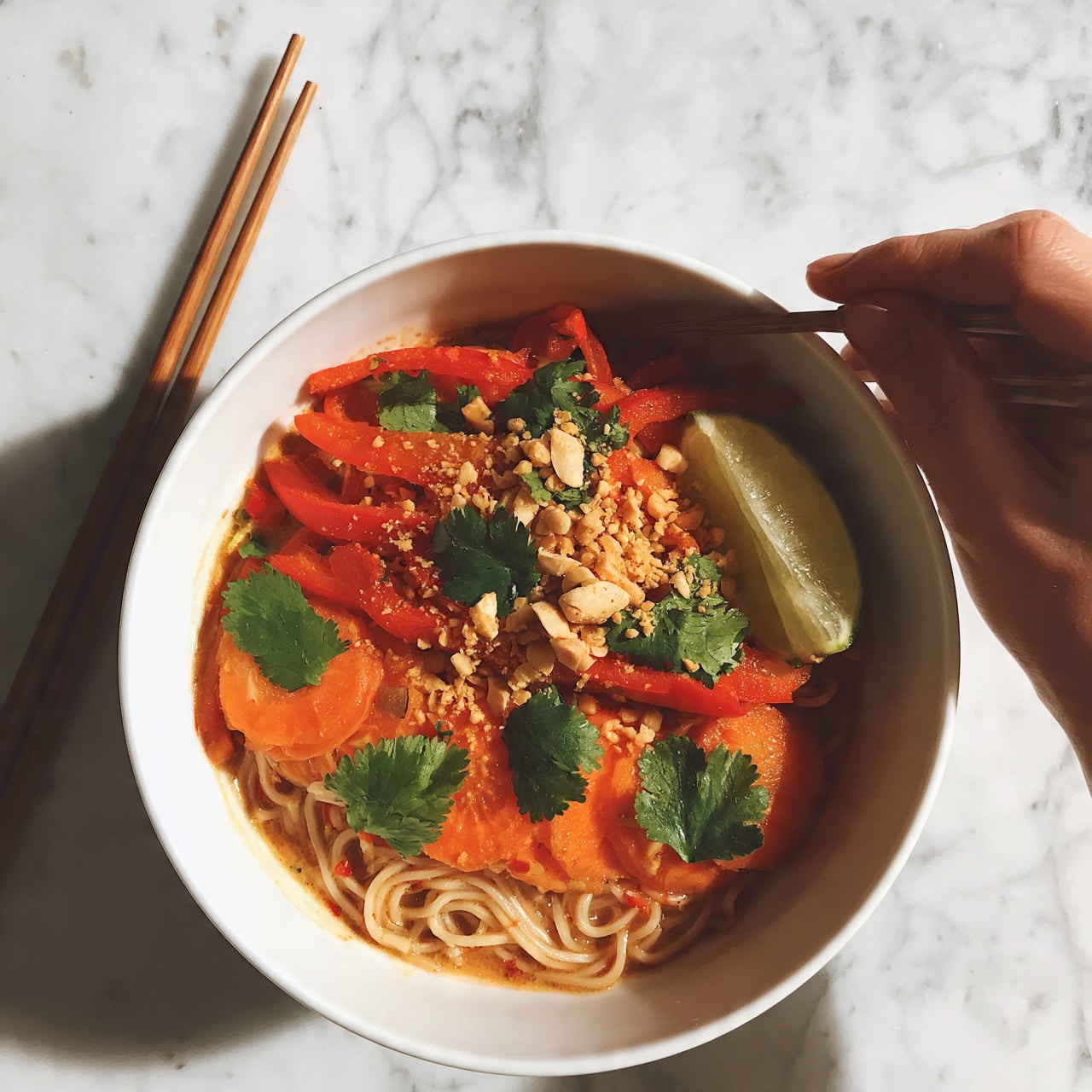 square image A white bowl filled with a colorful noodle dish sits on a white marbled surface. The dish has three main layers: at the bottom, thin noodles with a light brown sauce; on top, bright orange carrot slices, red bell pepper strips, and green cilantro leaves scattered evenly; the final layer includes crushed peanuts sprinkled over the vegetables, and a wedge of lime resting on one side. A pair of wooden chopsticks lies beside the bowl, angled slightly. A woman's hand is holding one chopstick gently near the bowl. Photo taken with an iphone --ar 4:5 --v 7