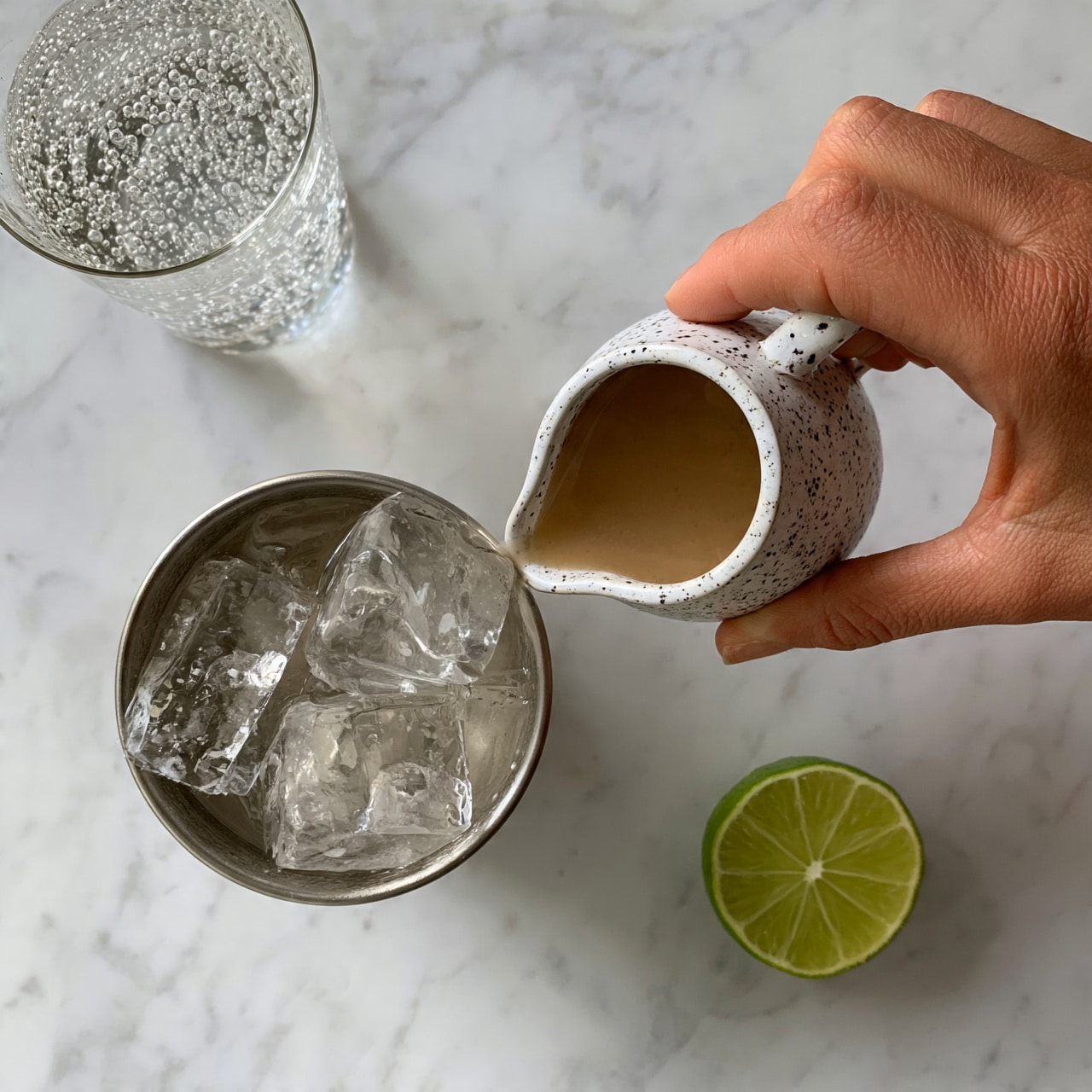 square image A woman's hand is pouring a light brown liquid from a small white ceramic pitcher with dark speckles into a metal cup filled with large, clear ice cubes. The cup is placed on a white marbled surface. Nearby, there is a glass of sparkling clear liquid with bubbles and a half lime showing a bright green outer skin and lighter green inside. photo taken with an iphone --ar 4:5 --v 7