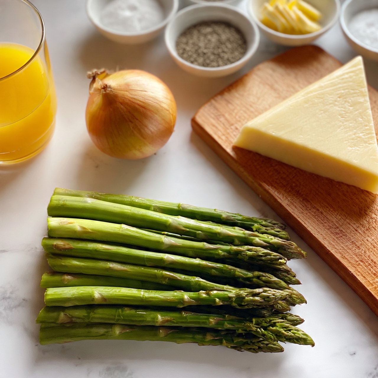 square image The image shows fresh asparagus spears neatly stacked in one layer on a white marbled surface near the bottom of the frame. Above them to the left sits a whole onion with golden brown skin. To the right of the onion is a wooden cutting board resting on the white marbled surface with a large, triangular piece of pale yellow cheese on it. Above the asparagus and onion, several small white bowls contain gathered ingredients: one with salt, one with black pepper, one with sliced butter, and one with a white powdery ingredient. To the far left is a clear glass filled with bright orange juice. The overall scene is well-lit and arranged cleanly. photo taken with an iphone --ar 4:5 --v 7 square image